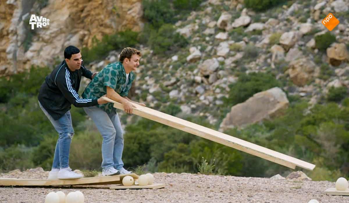 Twee mannen staan op een houten plank. Eén van de mannen houdt een houten plank vast. Er liggen grote eieren van de grond. Screenshot uit televisieprogramma Wie is de Mol.