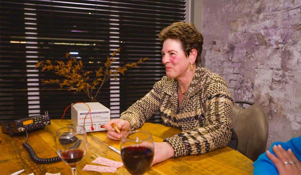 De zus van Derre zit met rode wangen en glazen wijn aan tafel bingo te spelen bij een oude radio in de serie Urk.