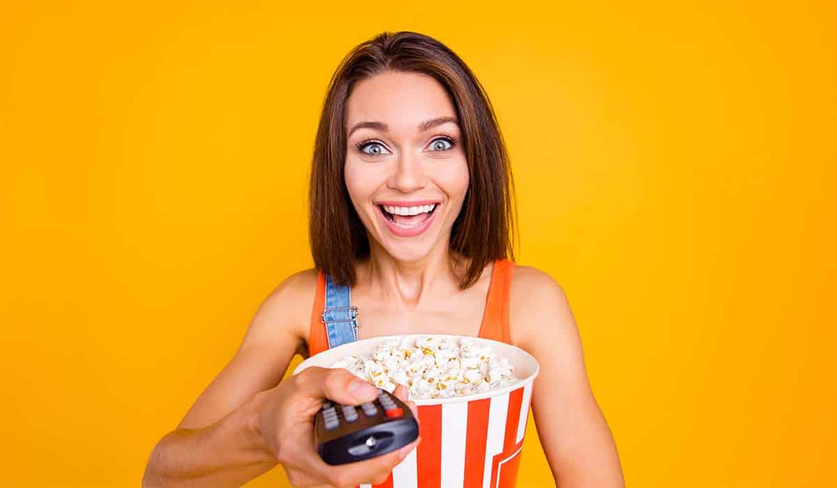 Lachende vrouw met popcorn en afstandsbediening in haar hand kijkt televisie. Credits: istock
