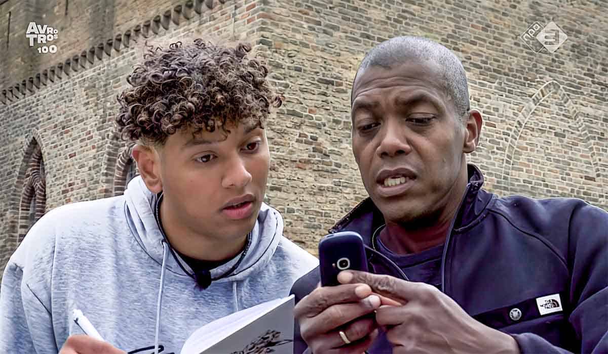 Hunted 2023 deelnemers Jesse en Prince, zoon en vader, staan voor een stenen muur en kijken op een mobiele telefoon en maken notities.